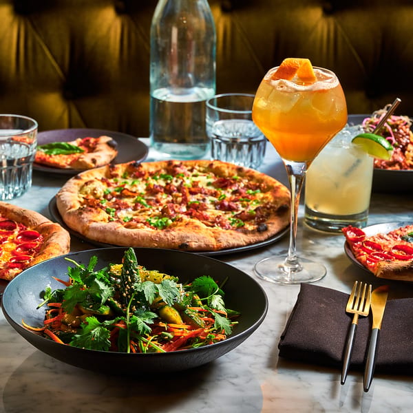 A marble table laden with pizzas, a salad, pasta, and cocktails in a dimly lit restaurant.