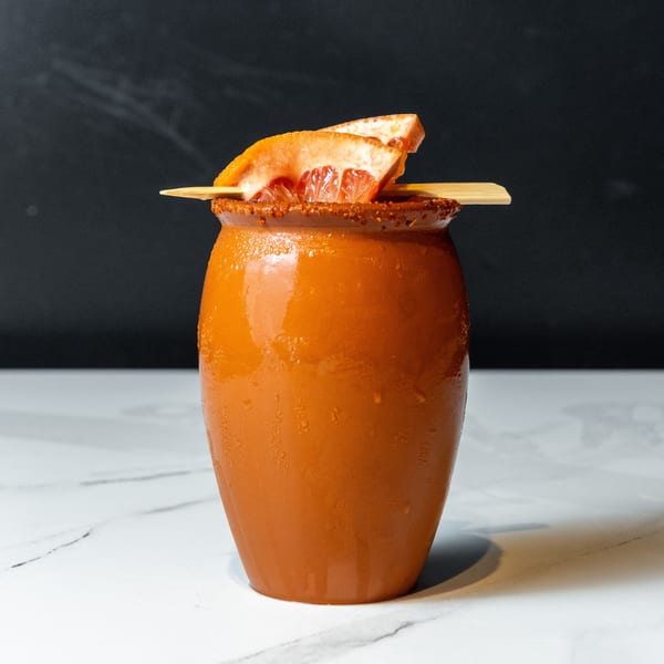A terracotta-colored cocktail in a ceramic jar, garnished with grapefruit slices and a chili-lime rim.