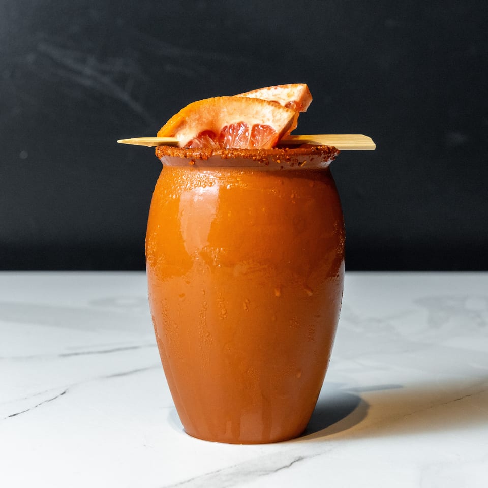 A terracotta-colored cocktail in a ceramic jar, garnished with grapefruit slices and a chili-lime rim.