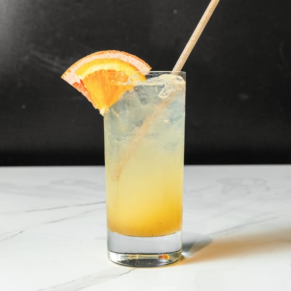 A tall glass of light yellow cocktail with ice, an orange slice, and a straw.