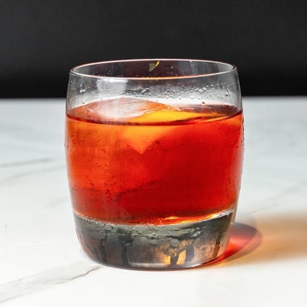 A vibrant red cocktail with ice and an orange slice in a glass on a white marble surface.