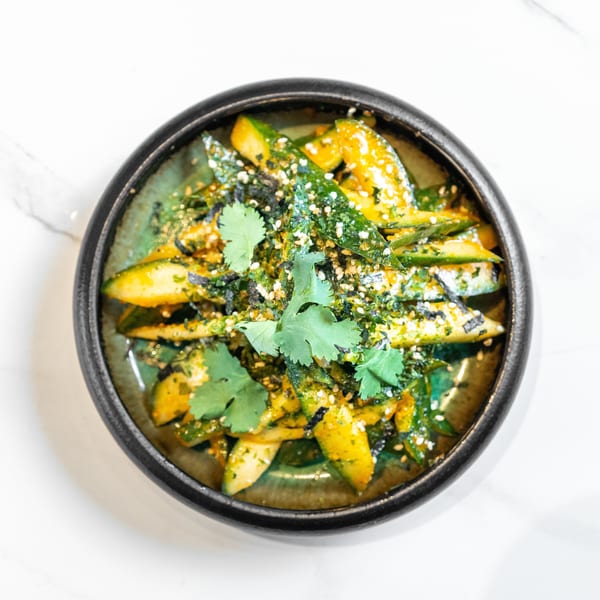 A vibrant green and yellow cucumber salad, garnished with cilantro, sesame seeds, and seaweed.