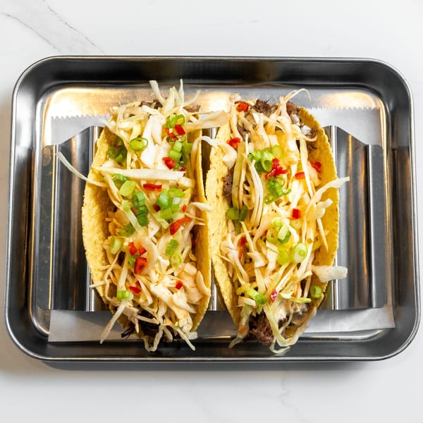 Two tacos with shredded meat, cabbage, green onions, and red peppers on a metal tray.