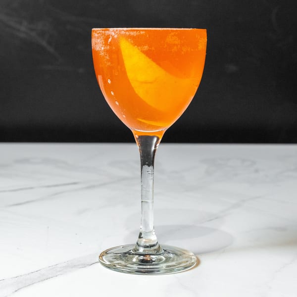 A vibrant orange cocktail in a coupe glass, garnished with an orange peel, on a marble surface.