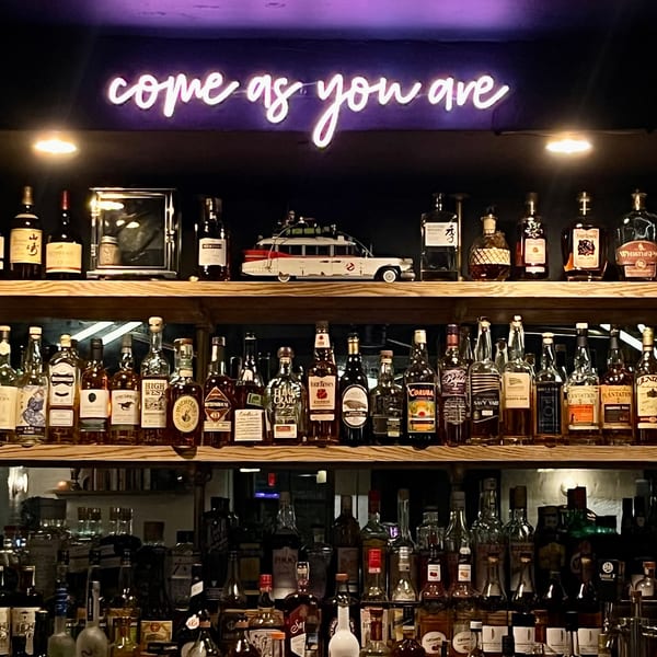 A neon sign above a bar reads "come as you are," illuminating shelves of liquor bottles.