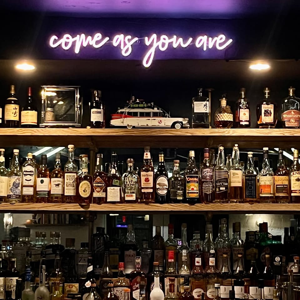 A neon sign above a bar reads "come as you are," illuminating shelves of liquor bottles.