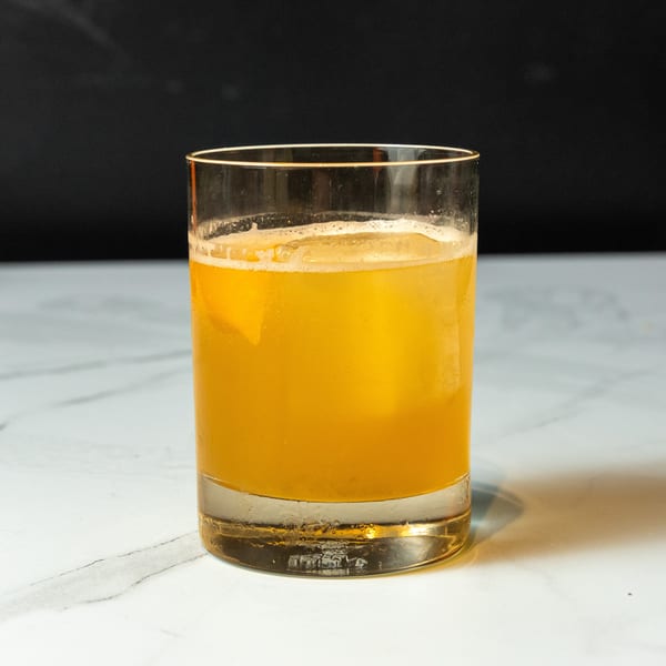 A glass of orange juice with ice and foam sits on a white marble surface.