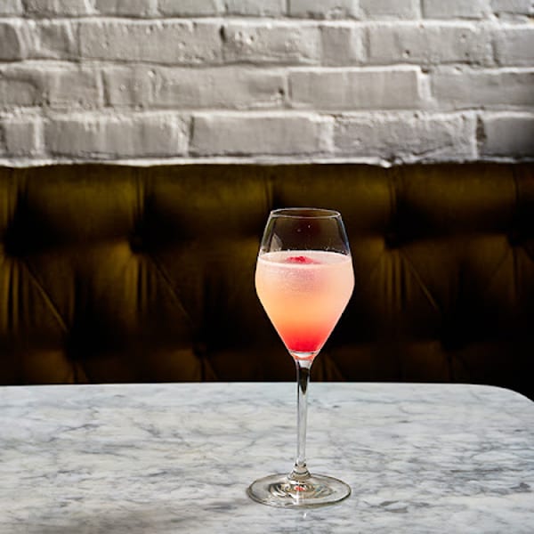 Pink and orange cocktail in a stemmed glass on a marble table with a brick wall background.
